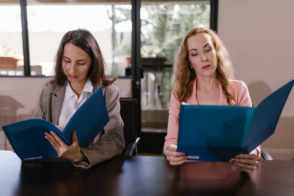 Two professional women sit at a table examining documents indoors, featuring teamwork and focus.